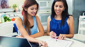 Two smiling young women sitting down at a kitchen table with a laptop and some academic papers.